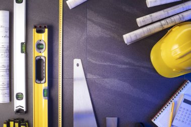 Müteahhit teması. Müteahhitin planları, defter ve alet çantası: sarı baret, iftiracı, gri fayansların arka planında testere ve yarda..