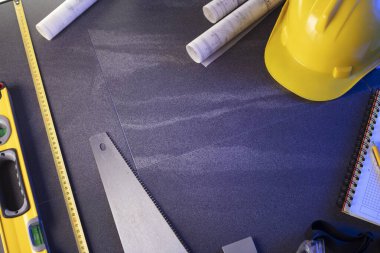 Müteahhit teması. Müteahhitin planları, defter ve alet çantası: sarı baret, iftiracı, gri fayansların arka planında testere ve yarda..