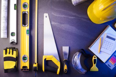 Müteahhit teması. Müteahhitin planları, defter ve alet çantası: sarı baret, iftiracı, gri fayansların arka planında testere ve yarda..