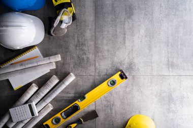 Müteahhit teması. Müteahhitin planları, defter ve alet çantası: sarı baret, iftiracı, gri fayansların arka planında testere ve yarda..