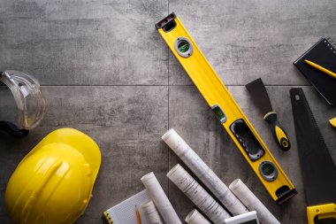 Müteahhit teması. Müteahhitin planları, defter ve alet çantası: sarı baret, iftiracı, gri fayansların arka planında testere ve yarda..
