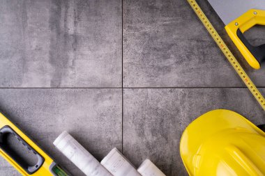 Müteahhit teması. Müteahhitin planları, defter ve alet çantası: sarı baret, iftiracı, gri fayansların arka planında testere ve yarda..
