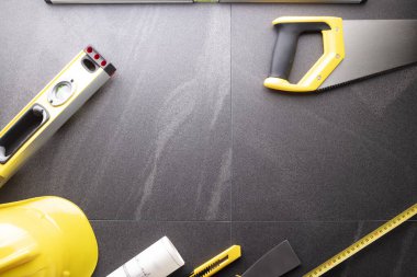 Müteahhit teması. Müteahhitin planları, defter ve alet çantası: sarı baret, iftiracı, gri fayansların arka planında testere ve yarda..