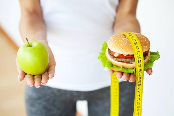 Joven chica atractiva está haciendo elección entre comida sana y dañina sosteniendo manzana y una hamburguesa en sus manos. — Foto de Stock