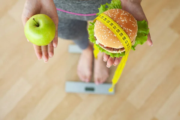 Ung attraktiv flicka gör valet mellan hälsosam och skadlig mat innehav äpple och en hamburgare i händerna. — Stockfoto