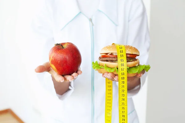 Giovane ragazza attraente sta facendo la scelta tra cibo sano e dannoso tenendo mela e un hamburger nelle sue mani. — Foto Stock