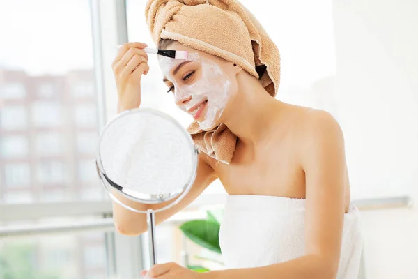 Mujer aplicando máscara en su cara y mirándose en el espejo . — Foto de Stock
