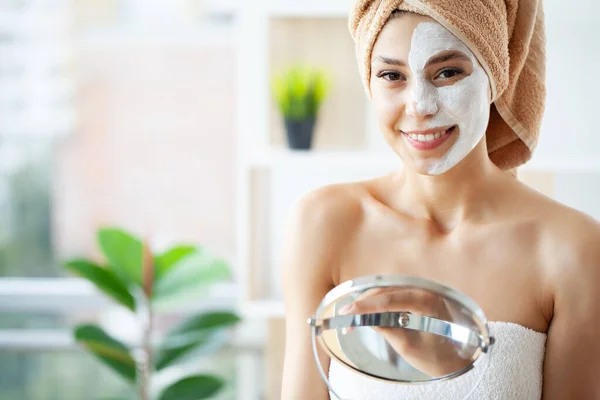 Beautiful young woman with towel wrapped around her head applying face mask at the bathroom — Stok Foto