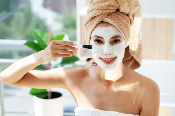 Beautiful young woman with towel wrapped around her head applying face mask at the bathroom — Photo