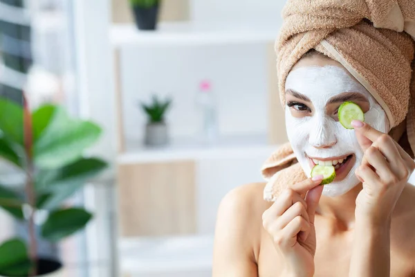 Hermosa joven con toalla envuelta alrededor de su cabeza aplicando mascarilla en el baño — Foto de Stock