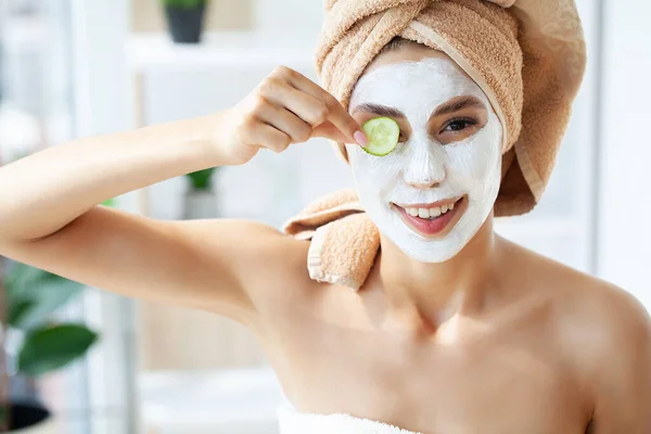 Wanita muda cantik dengan handuk melilit kepalanya mengenakan masker wajah di kamar mandi — Stok Foto