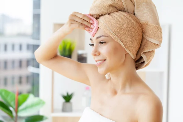 Glimlachende vrouw met gewikkeld in handdoek hoofd met behulp van gezicht spons — Stockfoto