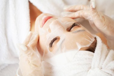 Kozmetik uzmanı spa salonundaki kadına çarşaf maskesi takıyor.