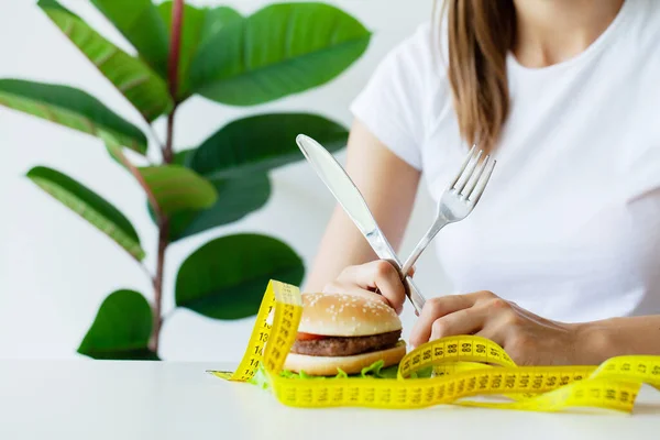 Mujer joven sosteniendo hamburguesa grasa dañina con cinta métrica amarilla — Foto de Stock