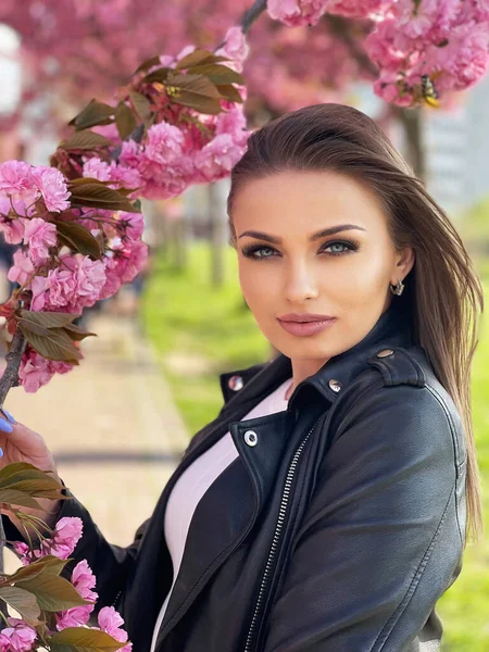 Schöne junge Frau in schwarzer Jacke steht im Frühling in der Farbe der Sakura-Blumen. — Stockfoto