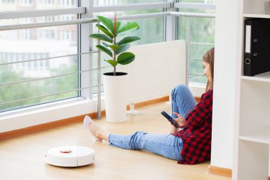 Beyaz robot süpürge zemini temizlerken kadın duvarın yanında oturup dinleniyor.