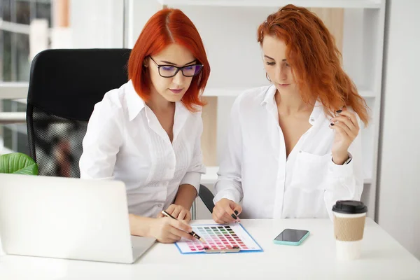 Two red-haired women work in the office at the table on a joint project.