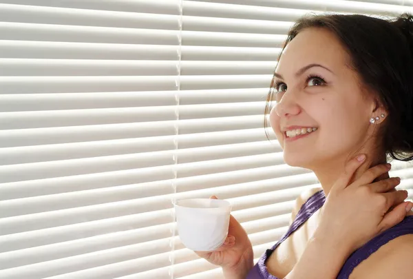 Ein gutes kaukasisches Mädchen, das mit einer Tasse am Fenster steht — Stockfoto