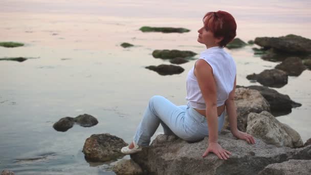 Вечером на берегу моря сидит гармоничная женщина — стоковое видео