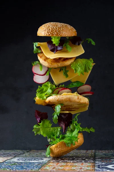 Pan de hamburguesa, doble chuleta de pollo con queso, lechuga, rúcula y rábano. Carne, verduras y pan. Ingredientes. Hamburguesa casera en un plato. Mesa hecha de azulejos de colores. —  Fotos de Stock