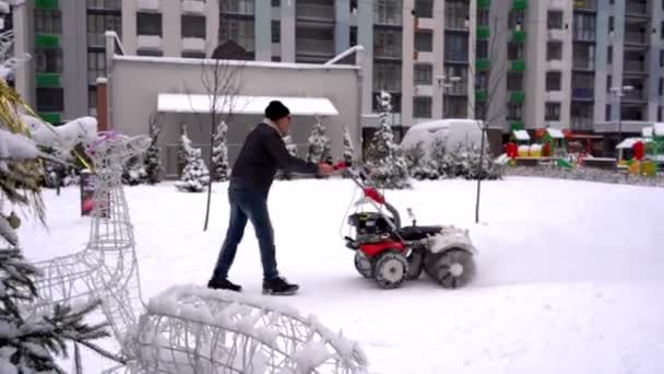 Clearing snow from paths in winter. Manual diesel tractor snow purifier. Grader. The area of the courtyard of a multi-storey building. Male janitor view from the back. — Stock video