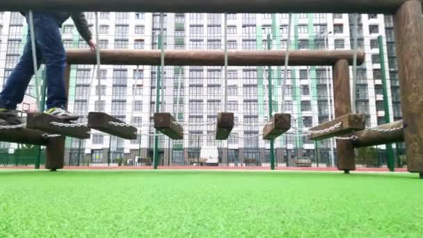 Swings on the new playground. The childs legs walk along wooden bars suspended on chains. Sports in the courtyard of a residential building. Go up the swinging stairs. The boy is playing. — стоковое видео