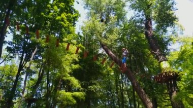 İp parkı. Miğferli bir genç çocuk asılmış halat merdivenlerle yürüyor. Jandarmalar ve emniyet kemerleri. Güvenlik. Yaz etkinliği. Spor. Ormandaki çocuk parkı..