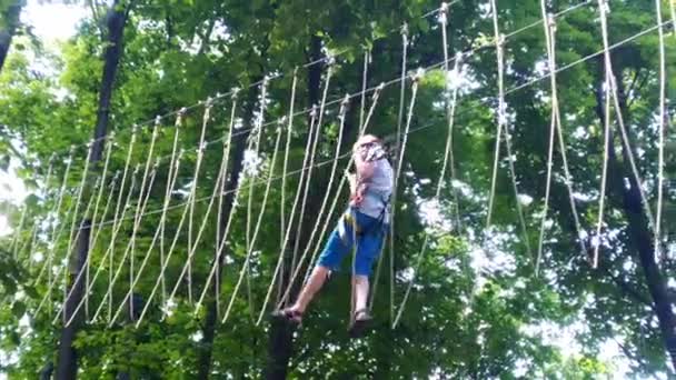 ロープパーク ヘルメットを着た少年10代の若者が吊り下げロープのはしごを歩く キャビナーと安全ストラップ 安全よ 夏のアクティビティ スポーツだ 森の中の自然の中の子供たちの遊び場 — ストック動画