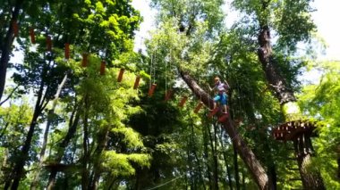 İp parkı. Miğferli bir genç çocuk asılmış halat merdivenlerle yürüyor. Jandarmalar ve emniyet kemerleri. Güvenlik. Yaz etkinliği. Spor. Ormandaki çocuk parkı..
