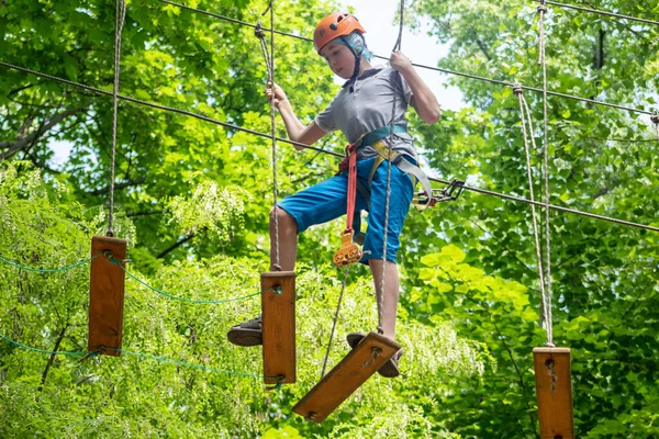 İp parkı. Miğferli bir genç çocuk asılmış halat merdivenlerle yürüyor. Jandarmalar ve emniyet kemerleri. Güvenlik. Yaz etkinliği. Spor. Ormandaki çocuk parkı..