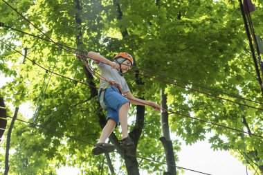 İp parkı. Miğferli bir genç çocuk asılmış halat merdivenlerle yürüyor. Jandarmalar ve emniyet kemerleri. Güvenlik. Yaz etkinliği. Spor. Ormandaki çocuk parkı..