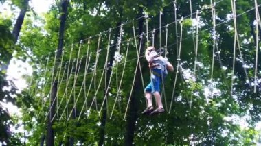 İp parkı. Miğferli bir genç çocuk asılmış halat merdivenlerle yürüyor. Jandarmalar ve emniyet kemerleri. Güvenlik. Yaz etkinliği. Spor. Ormandaki çocuk parkı..
