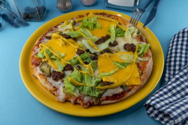 Etli hamburger, hardal, marul, peynir, sos. Mavi arka planda sarı tabakta yemek. Menü. Ev yapımı hamur işleri. Etli yemek. Masada yemek var..