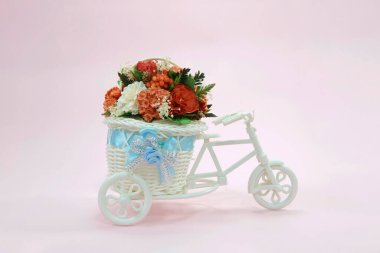 Küçük oyuncak beyaz bisiklet ve pembe arka planda bir sepet ve bir buket farklı yapay çiçek..