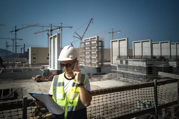 Caucasian civil engineer or construction worker in white hardhat uses his phone during inspection at construction site. Site engineer holds drawings calling on his mobile phone