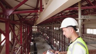 Site engineer dressed in white hardhat and protective vest makes notices to document on clipboard standing opposite industrial building. Foreman inspects truss structures of warehouse