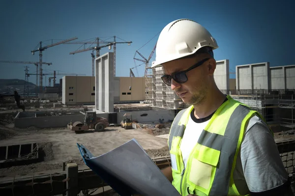 Caucasian civil engineer or construction worker dressed in white hardhat and protective vest looks at paperwork during inspection at construction site. Site engineer holds drawings on clipboard