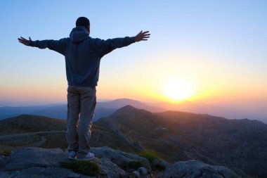 Male stands towards the sun on mountain slope with spread arms. Man meets the dawn with open arms. Sunrise on Nemrut Mount, Adiyaman province, Turkiye