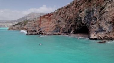 Aerial view of family supboarding to cave at secluded rocky beach. Mom and dad oaring on paddleboards with their sons down azure sea