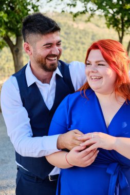 Turkish man happy embracing his caucasian plus size red headed fiancee, both laughing. Mixed race couple engagement. Middle eastern man and caucasian woman hugging.