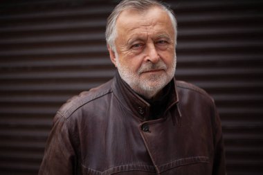 Gri sakallı bir adamın portresi. Bir sonbahar günü deri ceketli yaşlı bir adamın yakın çekimi. Arka Yuvarlanan Kapı