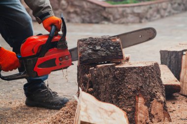 Elektrikli testere çalışıyor. Bir adam ağaç gövdesinin bir parçasını elektrikli testereyle kesiyor. Parça avludaki büyük bir ağaç kütüğünün üzerinde duruyor.