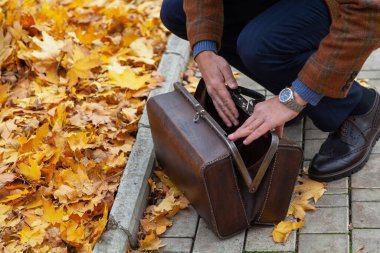 Kareli ceketli bir adam sonbahar parkının arka sokağında kahverengi bir halı çantası açıyor. Yukarıdan Görünüm
