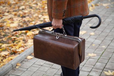 Şık bir ceketi olan beyefendi sonbahar öğleden sonra meydanın ortasında duruyor. Han 'ında eski bir çanta ve şemsiye kamışı tutuyor.