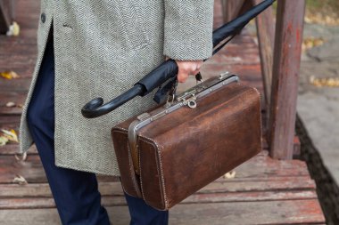 Köprüdeki beyefendi. Şık ayakkabılı ve gri paltolu bir adam sonbahar parkında ahşap bir köprü boyunca yürüyor. Elinde bir şemsiye ve valiz.