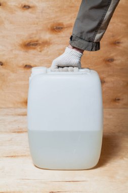 beyaz bir eldiven içinde beyaz bir plastik teneke kutu ile hafif bir kontrplak arka planı tutuyor. Konteynır likörle dolu.