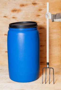 Çatal ve namlu. Mavi yuvarlak plastik bir konteynır açık renkli bir kontrplak arka planının karşısında duruyor. Tepesinde siyah bir kapak var. Sağ elinde beyaz bir eldiven, tahta sapında bir yaba tutuyor.