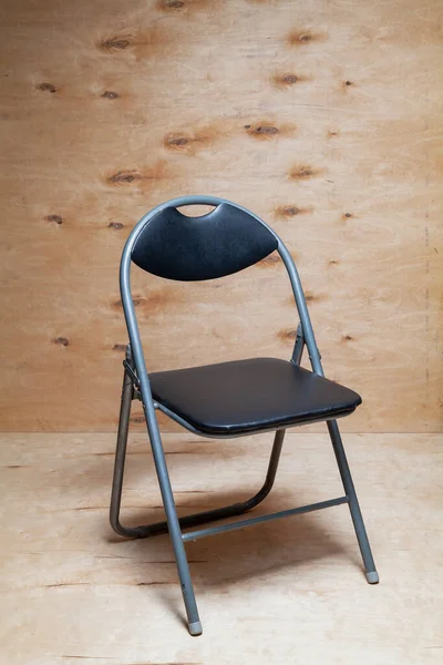 Art Nouveau folding chair stands against a background of light plywood. The chair has an arched back and a black sea
