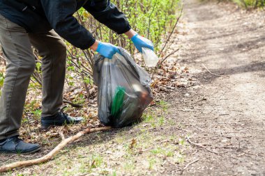 Doğayı çöpten kurtarıyoruz. Mavi lastik eldivenli ekolojist ön taraftaki siyah plastik torbada çöp topluyor.