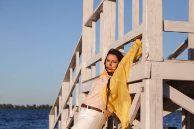 Güzel, sarı ceketli şişman bir kız kumlu bir sahilde beyaz ahşap bir yapının üzerinde yatıyor.